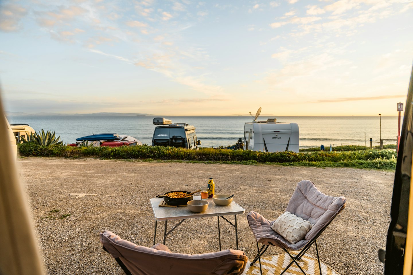 Blick vom Campervan auf einen Campingtisch mit Blick auf das Meer bei Sonnenuntergang.