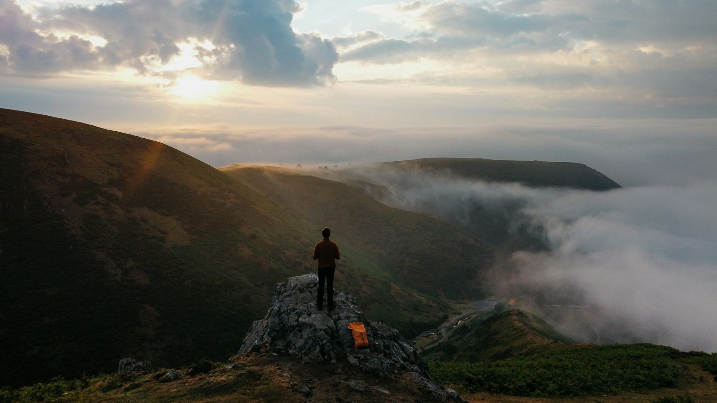 Wanderer steht auf einem felsigen Berggipfel mit Blick auf ein Flusstal.